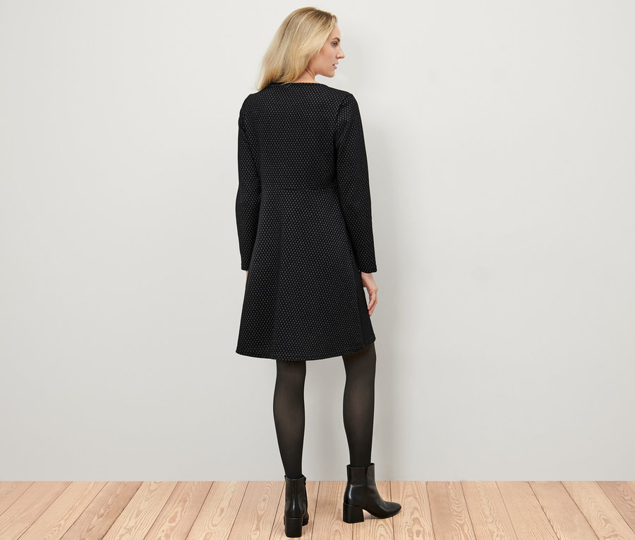 Blonde Frau steht mit dem Rücken zum Betrachter und trägt ein glitzerndes Jacquard-Kleid, eine schwarze Strumpfhose und Stiefel.