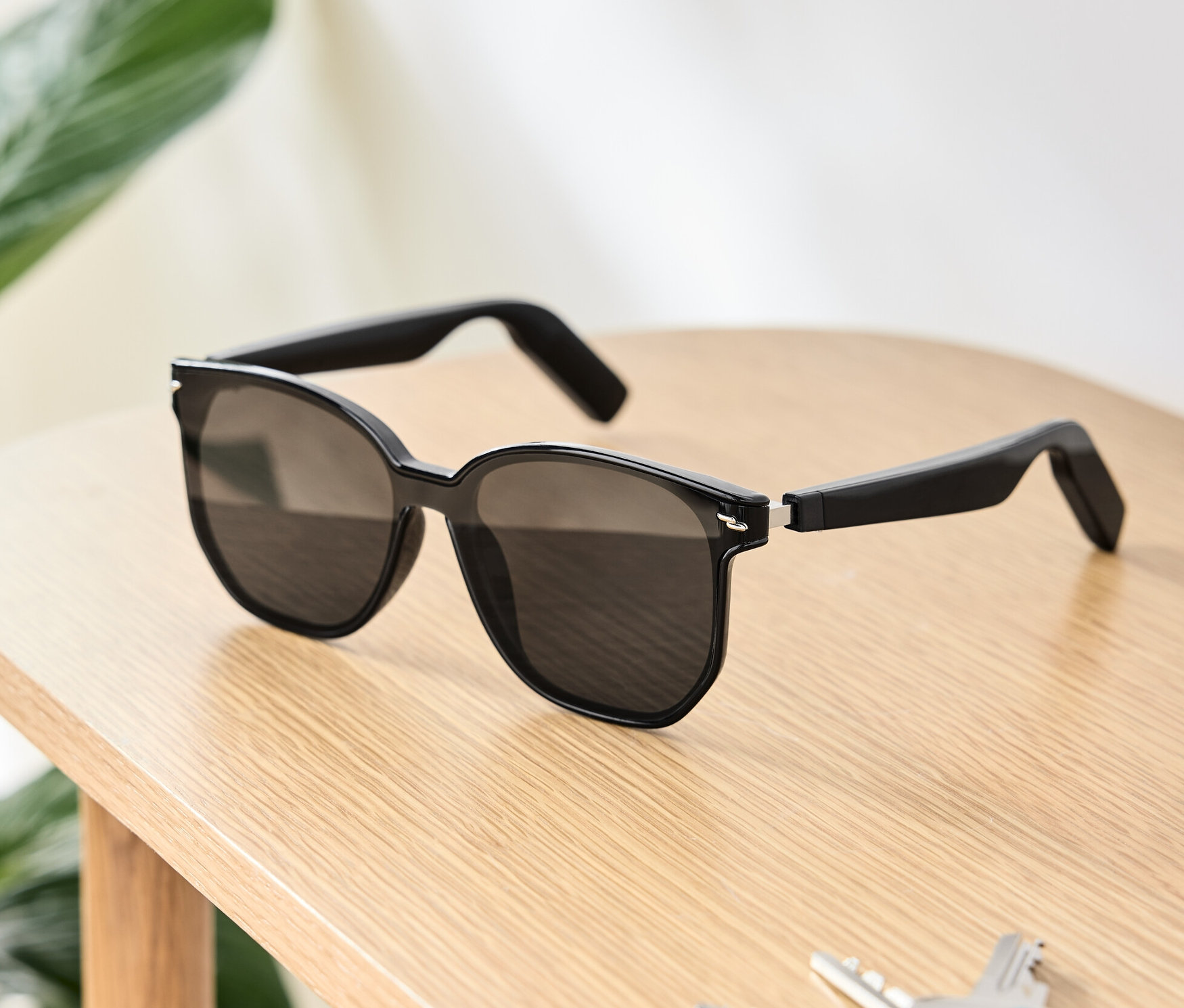 Eine schwarze Sonnenbrille mit Bluetooth®-Kopfhörern liegt auf einem Holztisch neben Schlüsseln und einer Geldbörse.