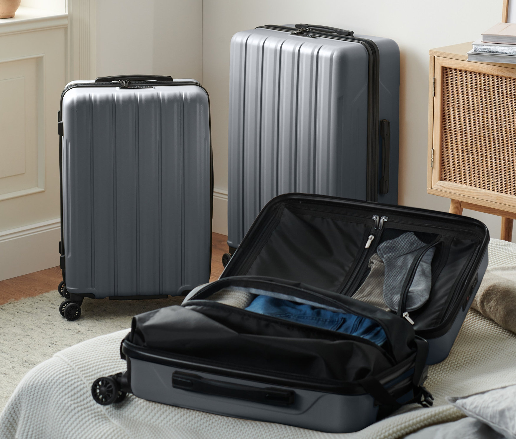 Drei graue Hartschalen-Koffer (klein, mittel und gross) stehen im Raum, einer davon geöffnet auf einem Bett.