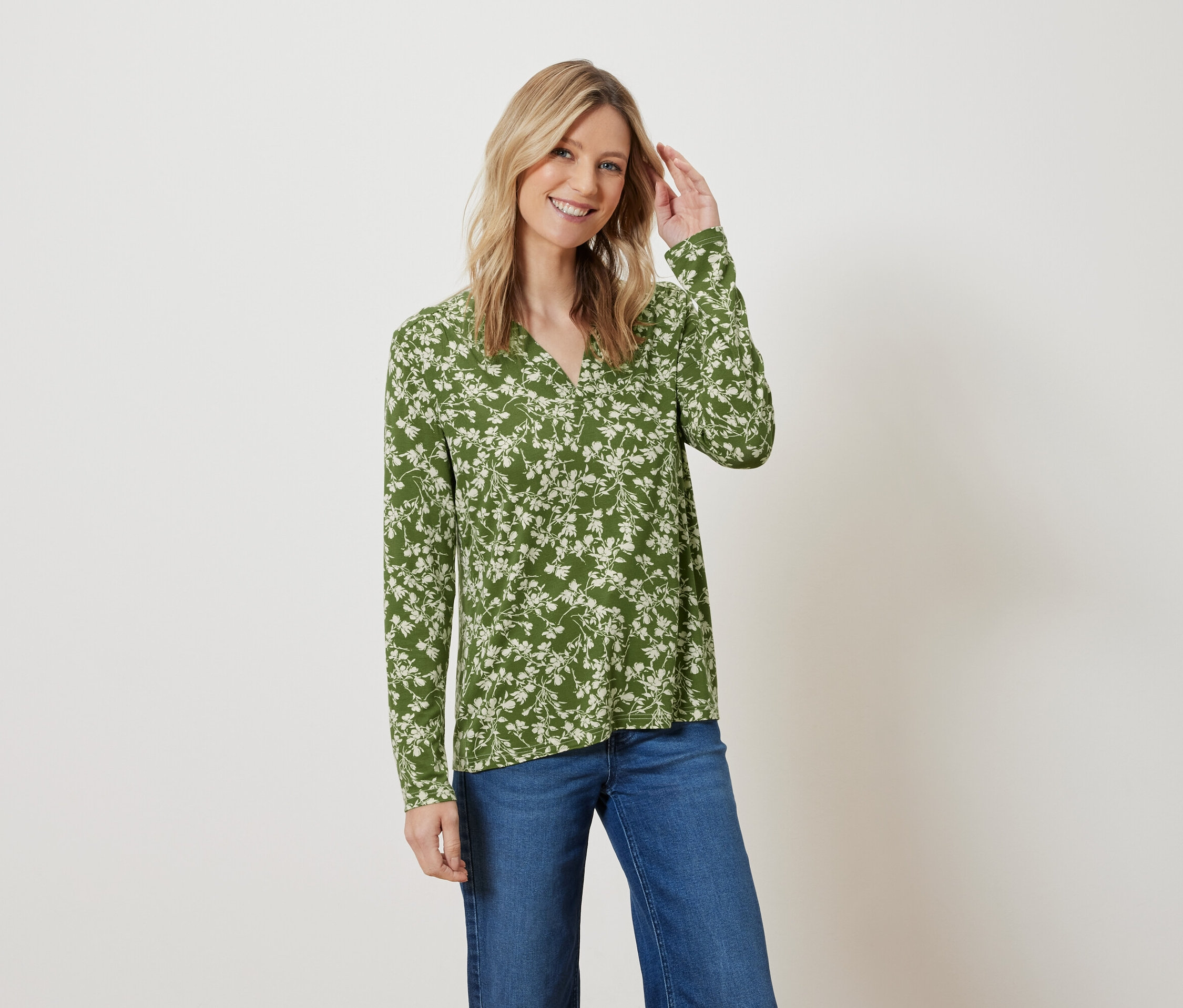 Frau mit blonden Haaren posiert in einer grünen Bluse mit Blumenmuster und blauen Jeans.