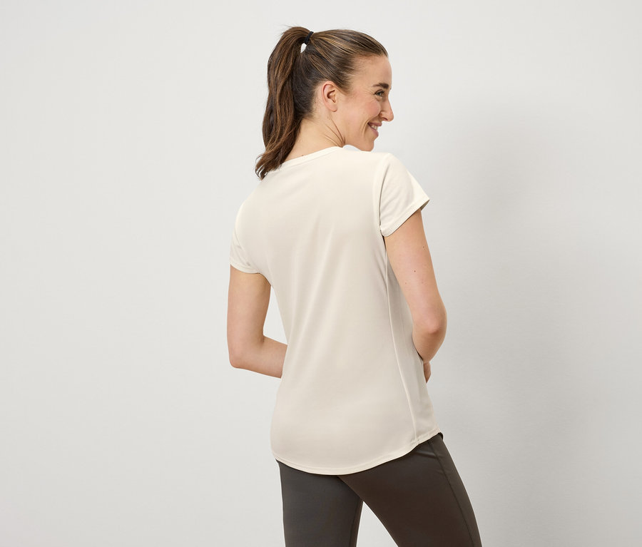 Eine Frau mit braunen, zu einem Pferdeschwanz zusammengebundenen Haaren lächelt und blickt nach links. Sie trägt ein cremefarbenes T-Shirt und braune Hosen.