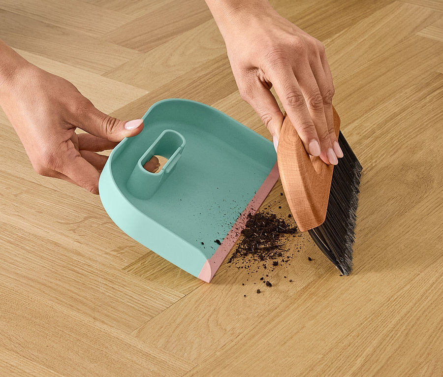 Frau kehrt Schmutz mit Handkehrer und Schaufel auf einem Holzboden zusammen.