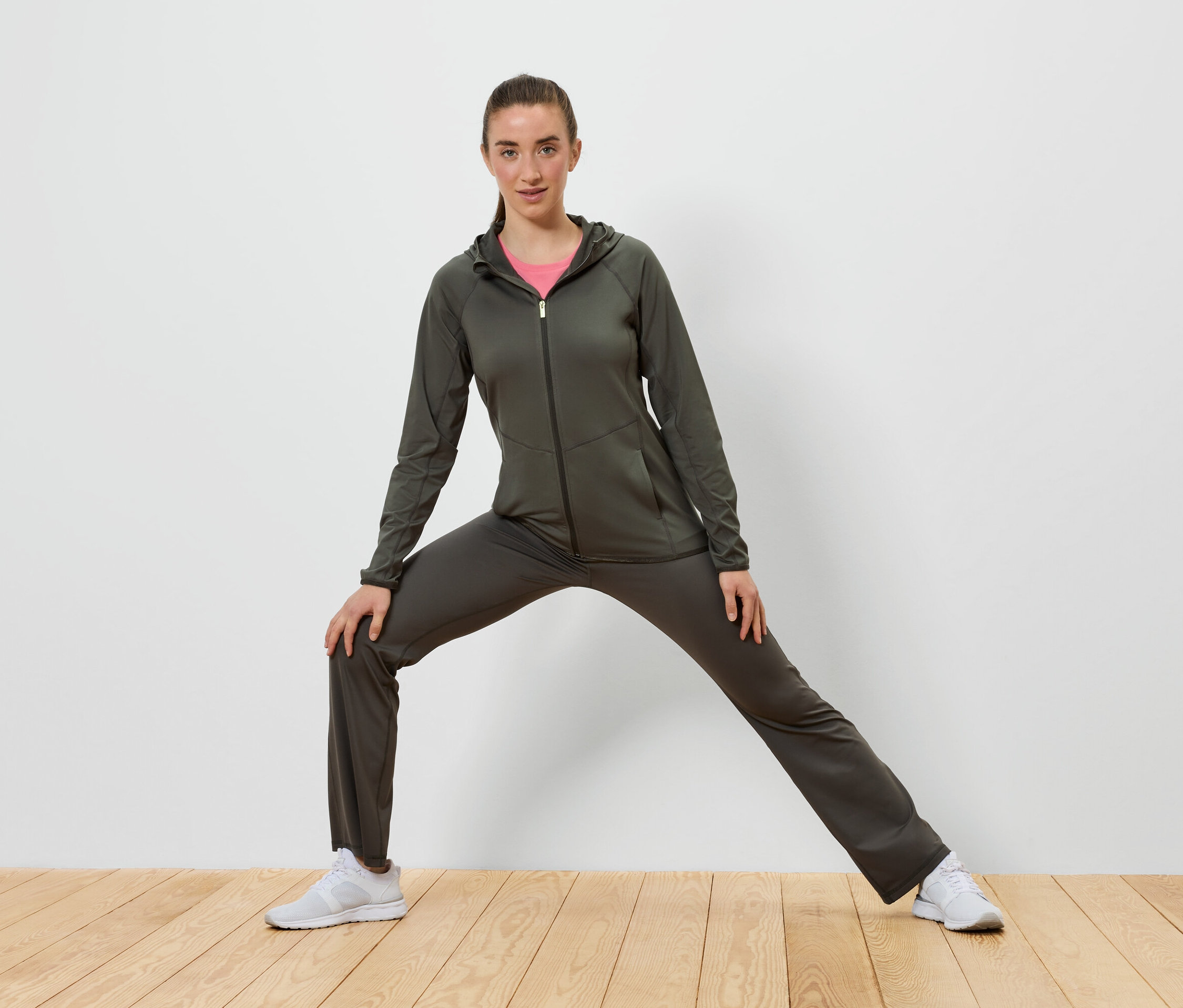 Frau in olivgrünem Trainingsanzug dehnt sich auf Holzboden vor einer weißen Wand.