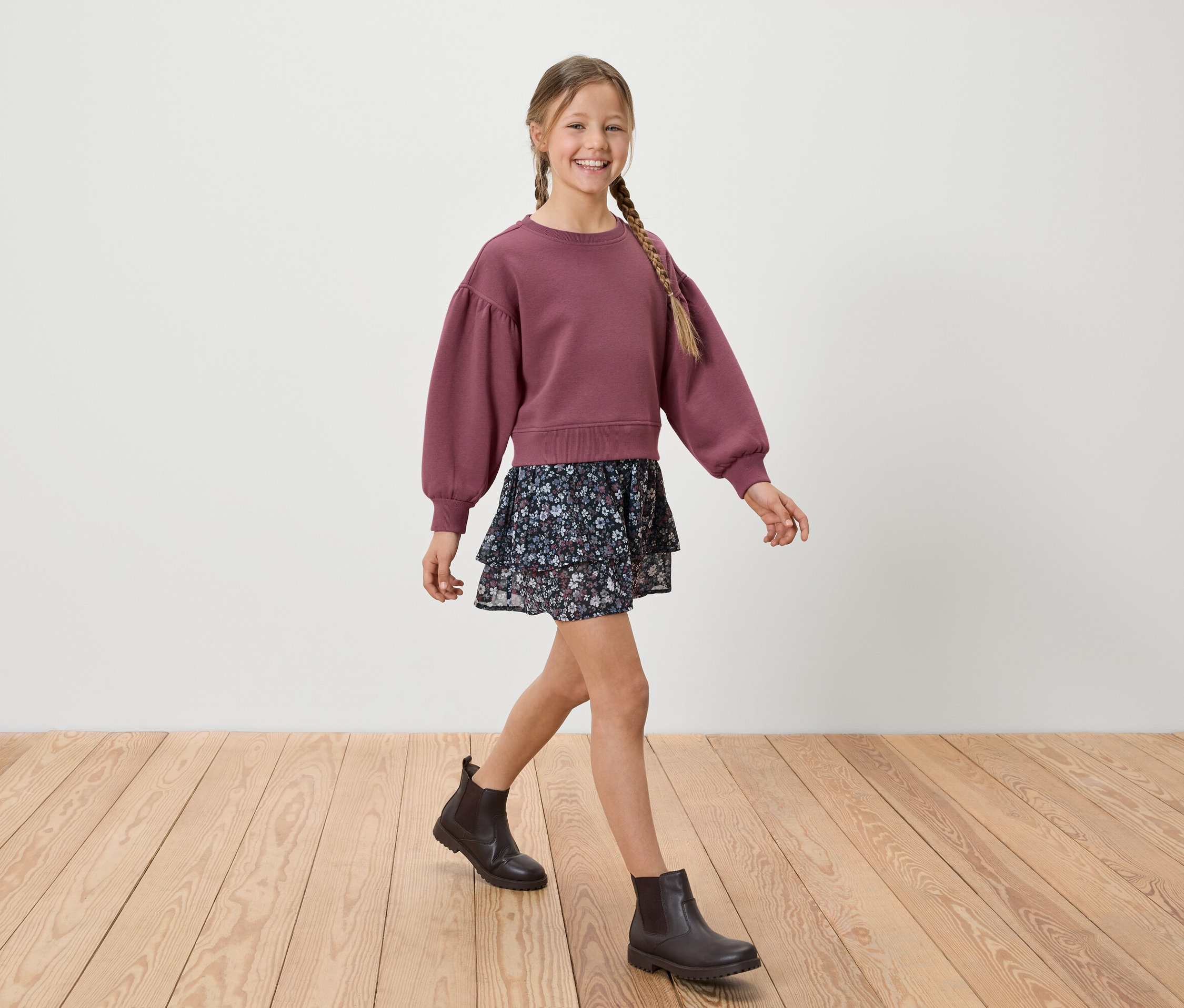 Ein lächelndes Mädchen mit Zopf geht auf einem Holzboden in einem bordeauxroten Sweatshirt, einem geblümten Rock und schwarzen Stiefeln.