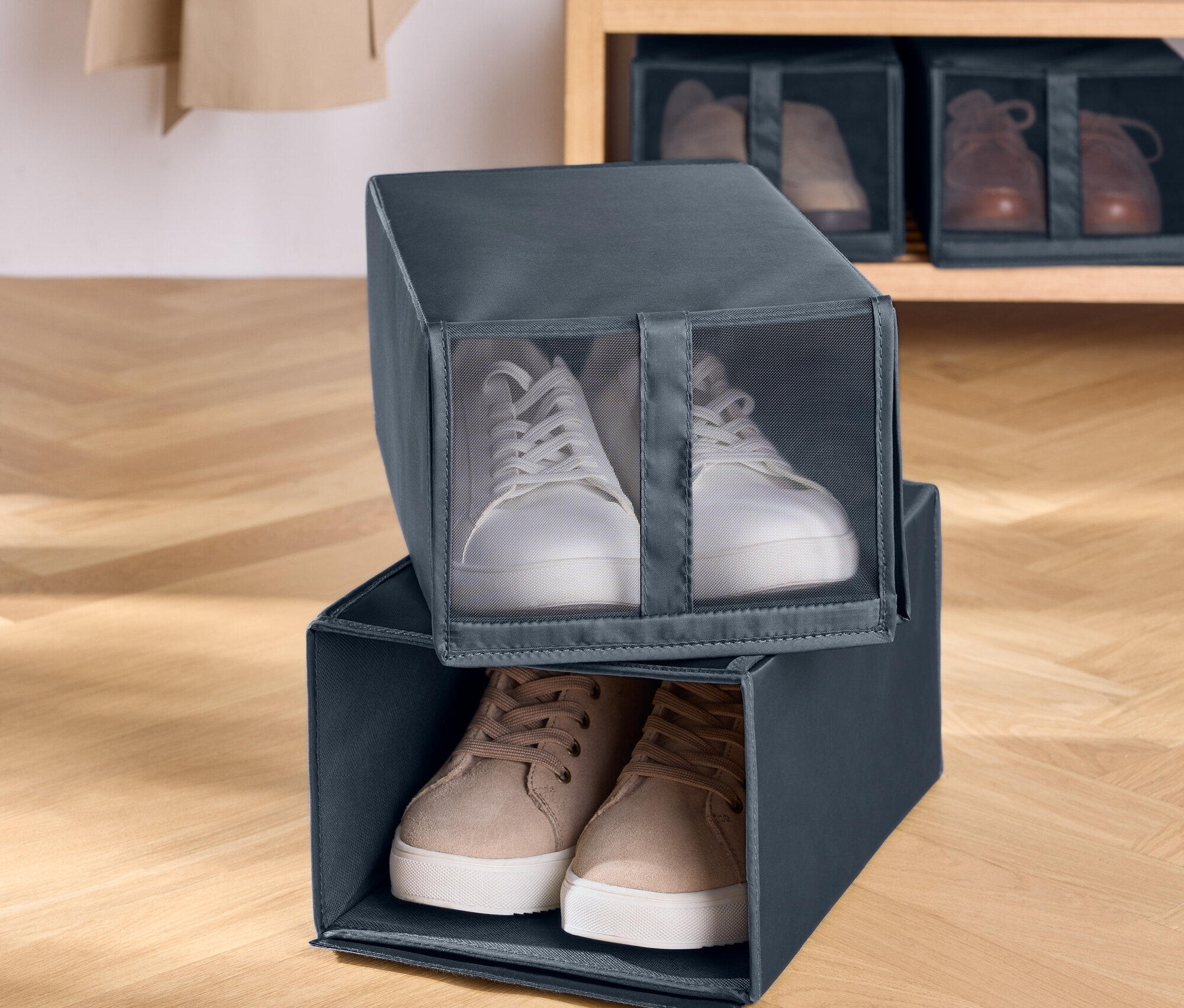 Zwei graue Stoff-Schuhboxen sind übereinander gestapelt. In der oberen Box befinden sich weiße Turnschuhe und in der unteren Box braune Turnschuhe. Im Hintergrund befindet sich ein Holzregal mit Schuhen.