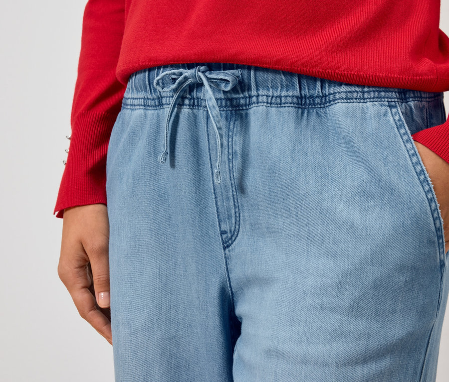 Teilansicht einer Person mit rotem Pullover und hellblauen Jeans mit der Hand in der Tasche.