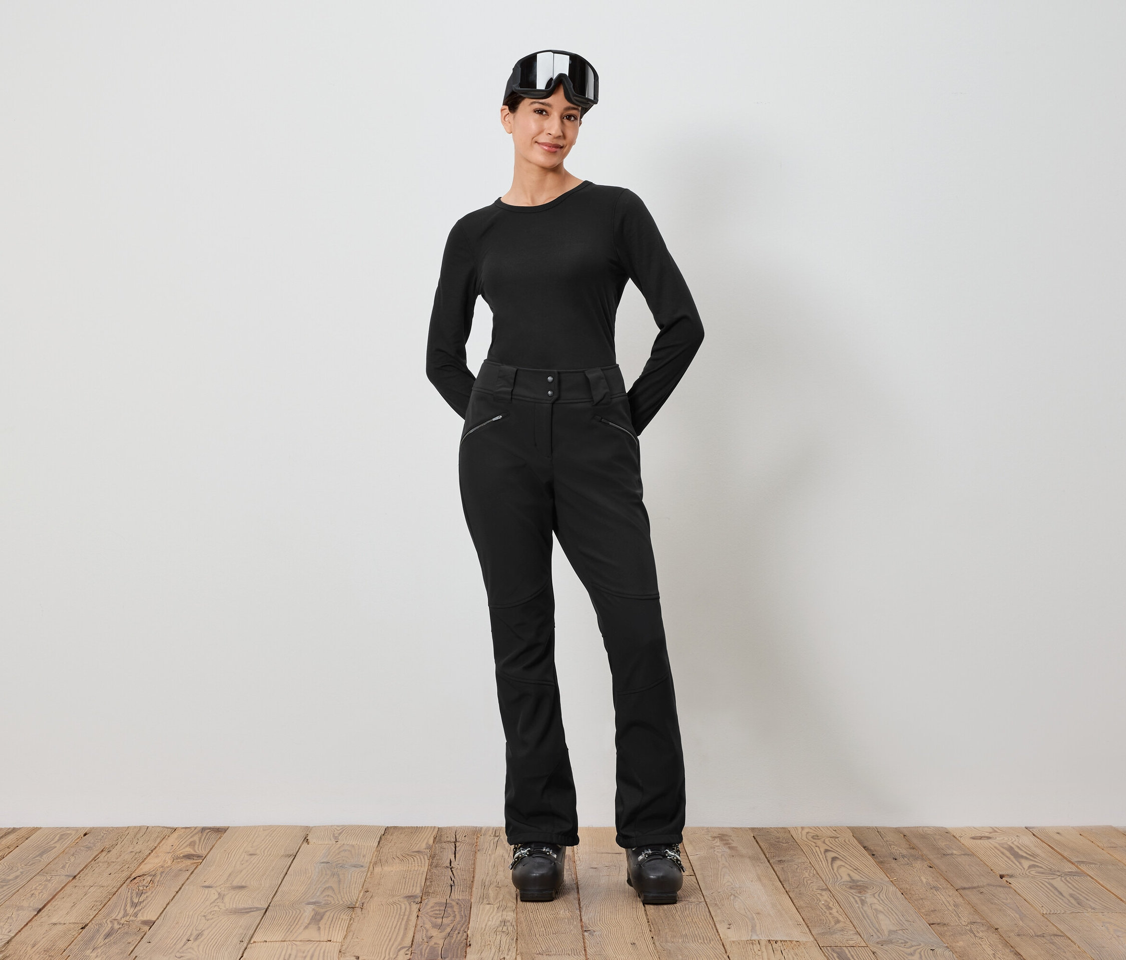 Frau in schwarzer Skibekleidung und Skibrille auf dem Kopf steht vor einer weißen Wand.