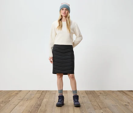 Eine Frau steht auf einem Holzboden und trägt einen cremefarbenen Pullover, einen schwarzen Rock, Stiefel und eine Strickmütze.