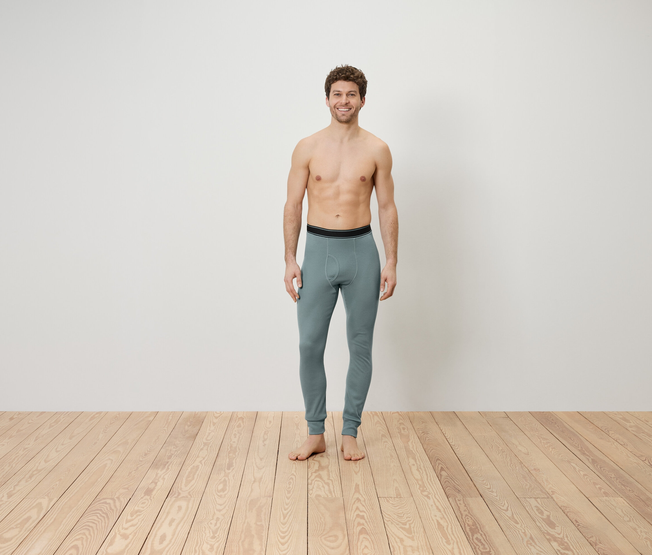 Ein Mann mit freiem Oberkörper steht auf einem Holzboden und trägt graue Leggings.