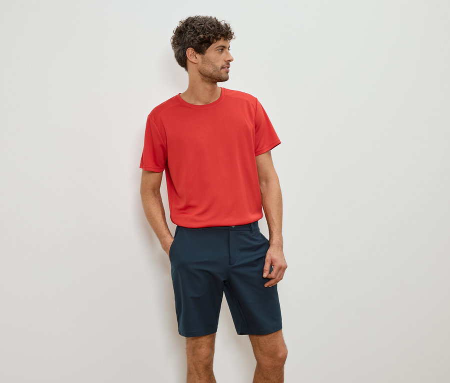 Mann mit rotem T-Shirt und blauen Shorts, die Hände in den Taschen.