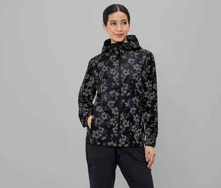 Frau mit Kapuze und Blumenmusterjacke, die in ihre schwarze Hose greift, vor grauem Hintergrund.