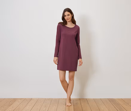 Eine Frau steht barfuß vor einer weißen Wand auf einem Holzboden und trägt ein weinrotes, langärmeliges Kleid.