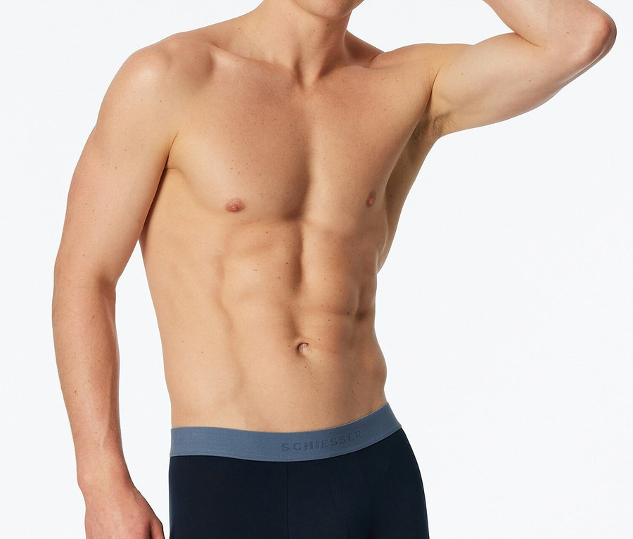 Mann ohne Hemd posiert in marineblauer Boxershorts mit blauem Bund vor einem weißen Hintergrund.