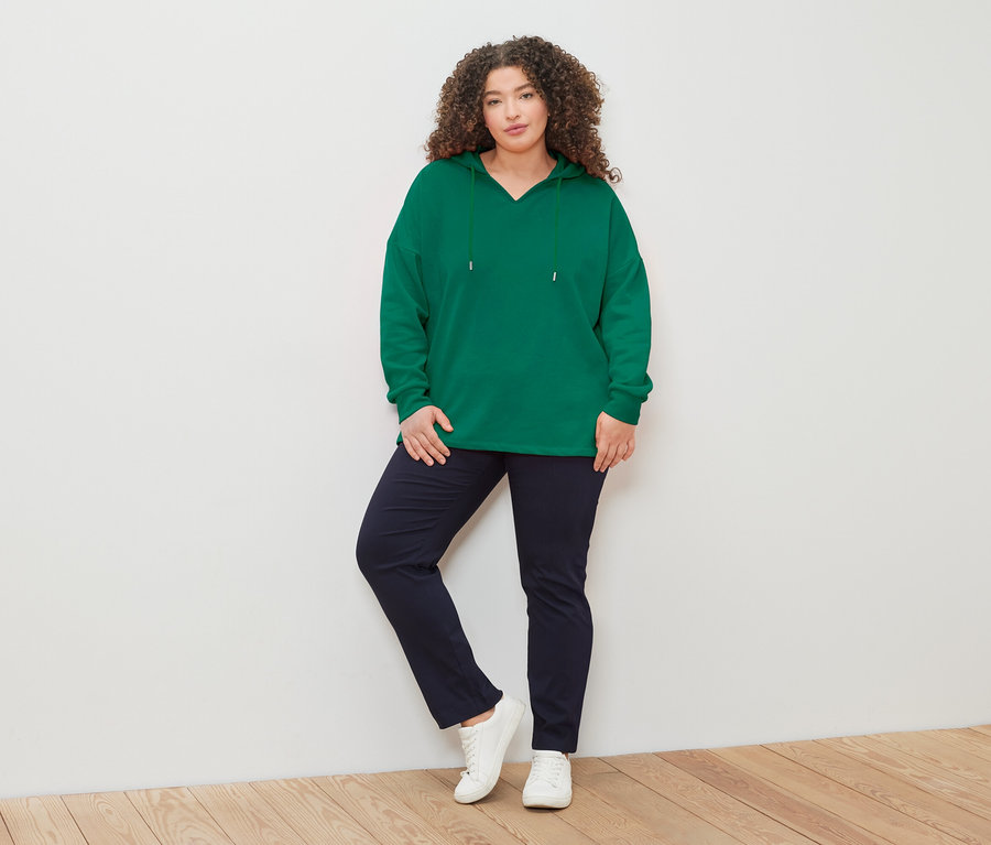 Frau mit lockigem Haar, trägt einen grünen Kapuzenpullover, blaue Hose und weiße Turnschuhe, posiert vor einer weißen Wand.