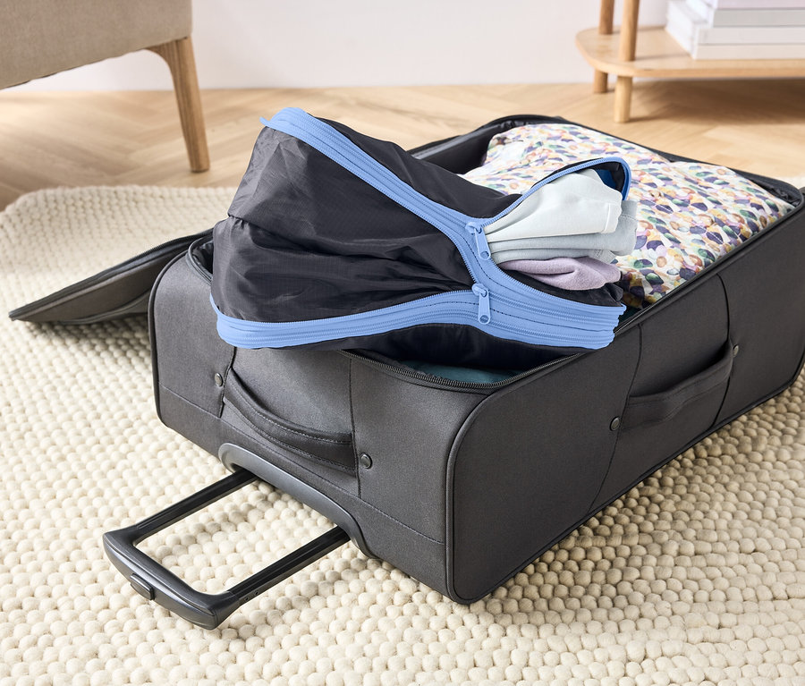 Offener, grauer Koffer auf einem Teppich, gefüllt mit Kleidung und einem schwarzen Packwürfel mit blauem Reißverschluss.