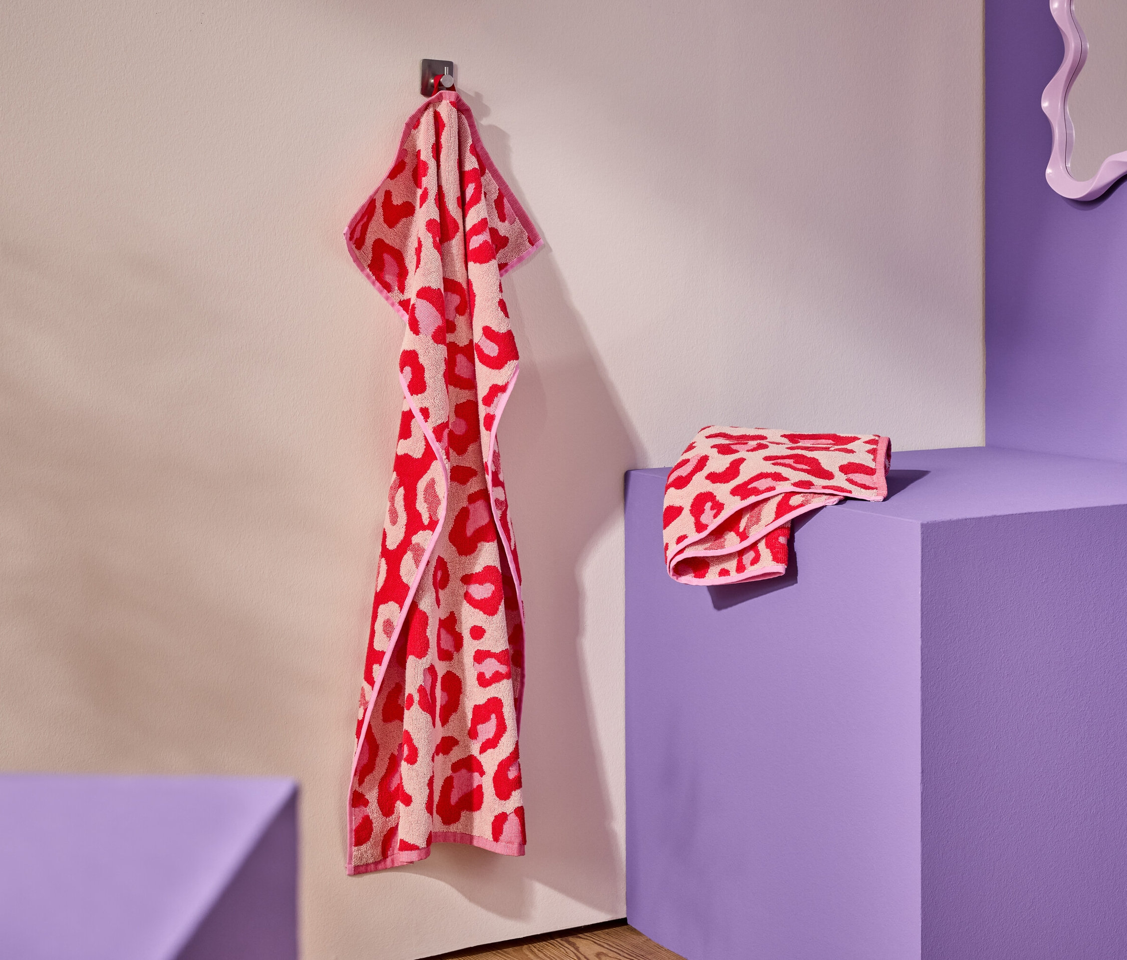 Ein rot-pinkes Handtuch mit Leopardenmuster hängt an einem Haken. Ein gefaltetes Handtuch liegt auf einem lila Podest.