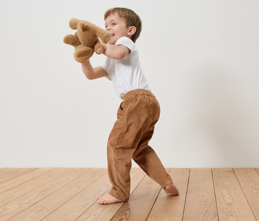 Ein Junge in Cordhose und T-Shirt spielt barfuß mit einem Teddybär vor einer weißen Wand.