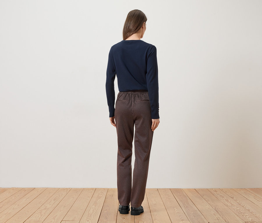 Eine Frau von hinten in marineblauem Oberteil und gemusterter Hose steht auf einem Holzboden vor einer weißen Wand.