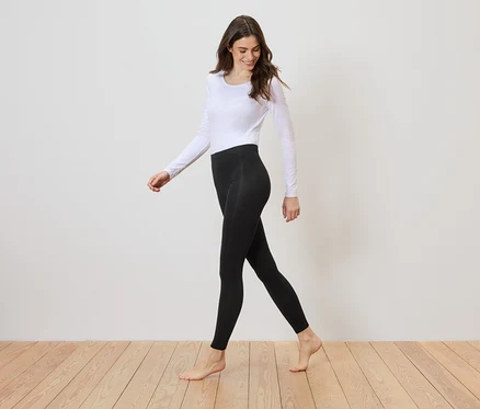 Barfüßige Frau mit langärmligem, weißen Oberteil und schwarzer Leggings geht auf einem Holzboden vor einer weißen Wand entlang.