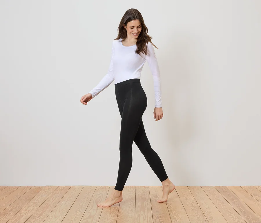 Barfüßige Frau mit langärmligem, weißen Oberteil und schwarzer Leggings geht auf einem Holzboden vor einer weißen Wand entlang.