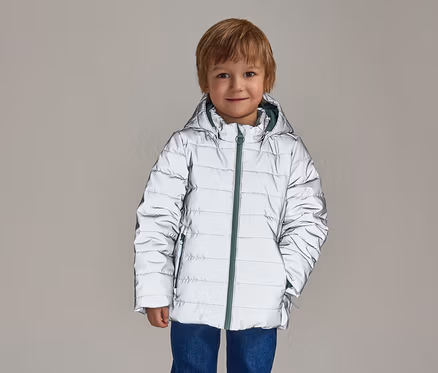Ein lächelnder Junge mit blonden Haaren trägt eine reflektierende, silberne Pufferjacke mit Kapuze und grünem Reißverschluss über blauen Jeans.