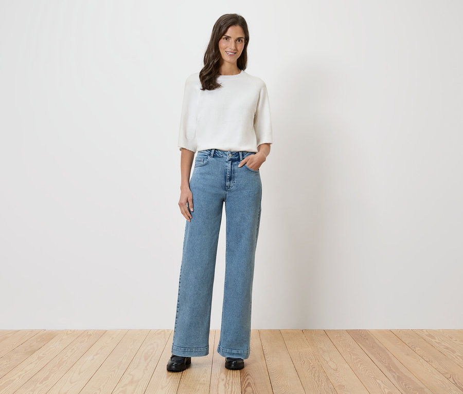 Eine Frau steht vor einer weißen Wand und trägt eine weiße Bluse und weite Jeans.