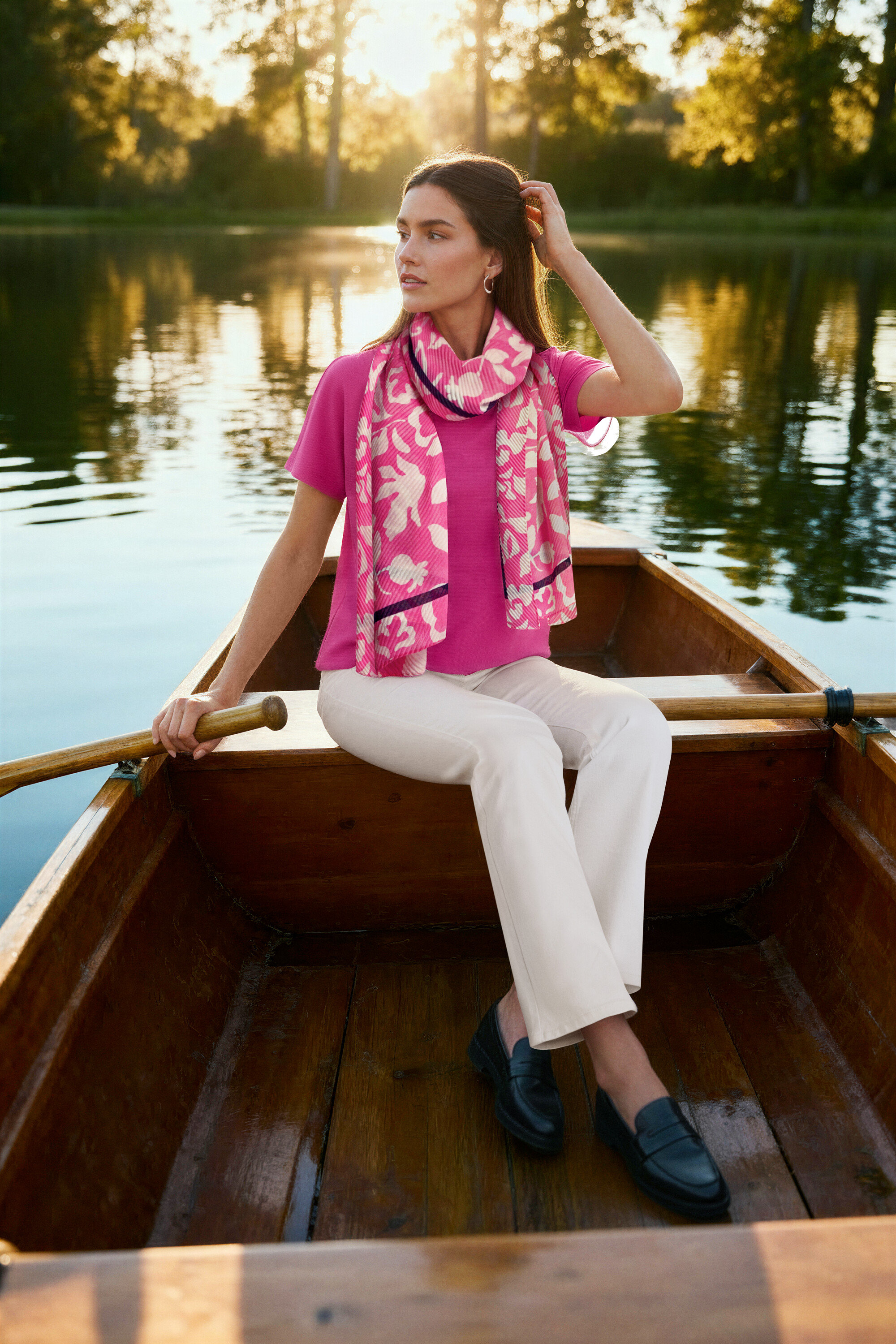 Frau sitzt in einem Ruderboot, trägt eine pinkfarbene Bluse, cremeweiße Bootcut Jeans ‒ Fit »Lou«, Leder-Loafer und Creolen Wave.