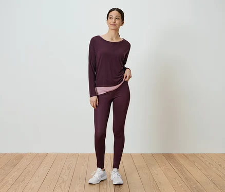 Frau steht und trägt ein langärmliges, auberginefarbenes Hemd und auberginefarbene Leggings.