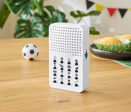 Weißes Gerät mit Knöpfen auf einem Holztisch, mit einem kleinen Fußball, Muffins und Partydekorationen im Hintergrund.
