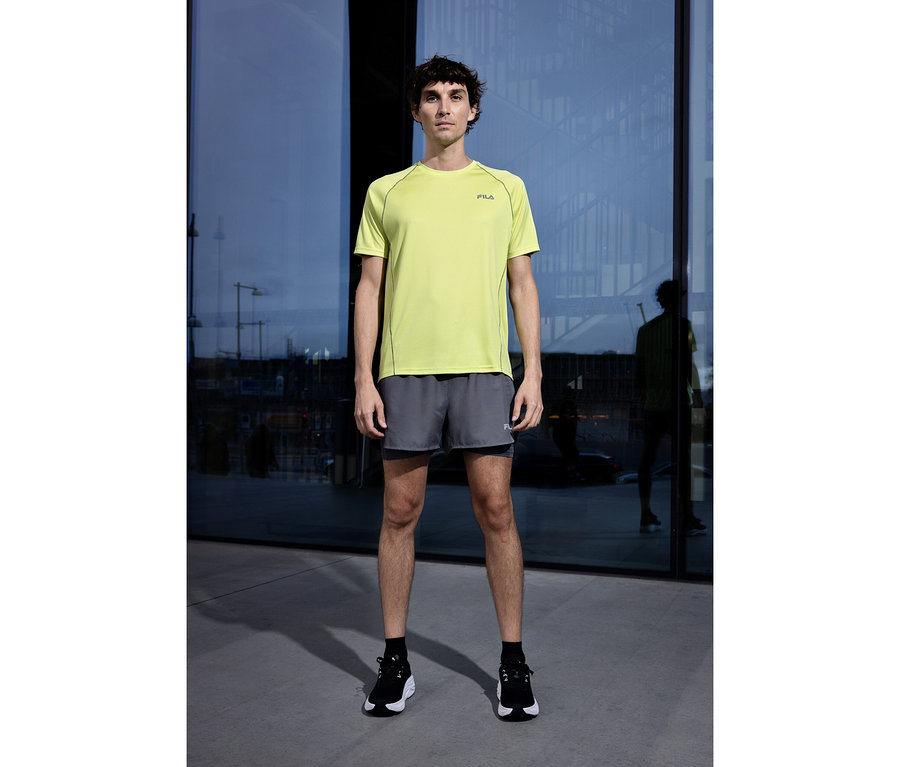 Mann steht vor Glasfassade, trägt ein hellgrünes FILA Herren-Funktionsshirt, graue Shorts und schwarze Sportschuhe.