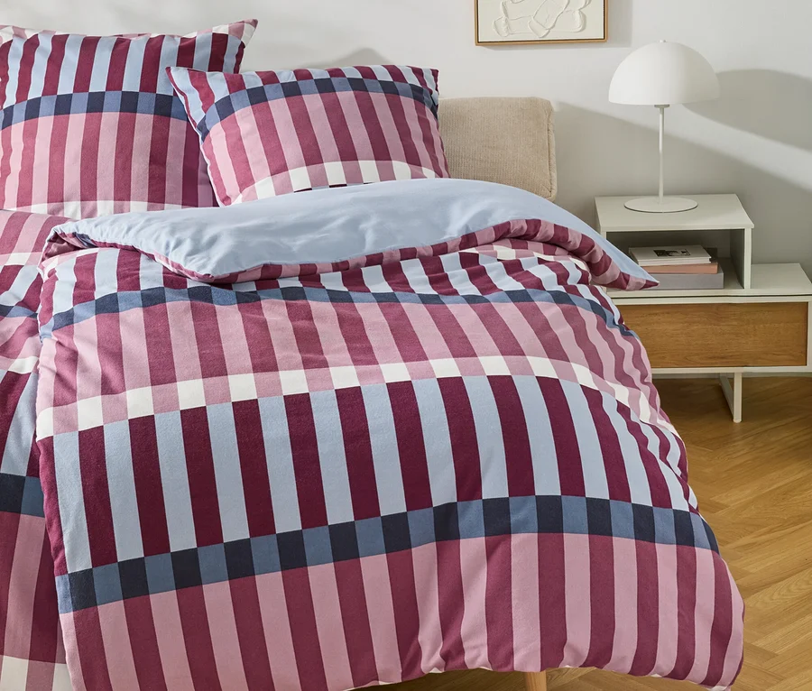 Ein Bett mit einem karierten Bettbezug in Rosa, Marineblau und Hellblau, dazu passende Kissen. Rechts daneben steht ein weißer Nachttisch mit einer weißen Lampe und Büchern. An der Wand hängt ein abstraktes weißes Reliefbild.