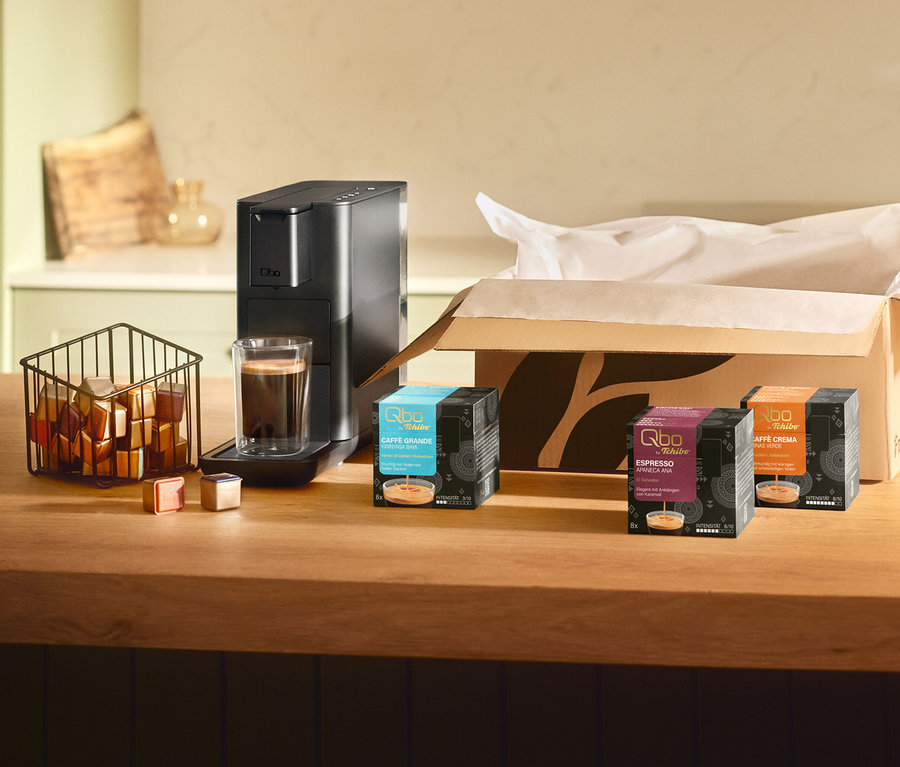 Schwarze Qbo Iconic, Dark Chrome Kaffeemaschine mit Kaffee in Tasse, Kapseln in Korb. Daneben drei Qbo Kaffeekapselpackungen: Qbo Caffè Grande KIRINYAGA BAYA, Qbo Espresso APANECA ANA, Qbo Caffè MINAS VERDE auf Holztheke.