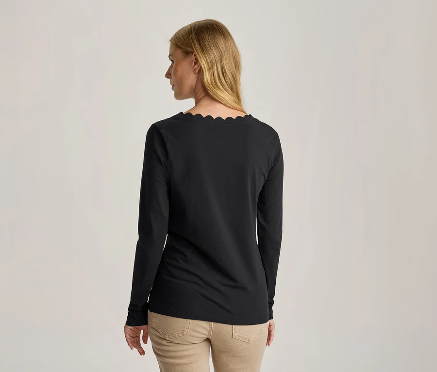 Frau von hinten, leicht gedreht, trägt ein schwarzes Langarmshirt mit Wellenausschnitt und beigefarbene Hose.