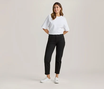 Stehende Frau in weißem Oversize-T-Shirt, schwarzer Cropped-Jeans und weißen Sneakern, mit Händen hinter dem Rücken lächelnd.