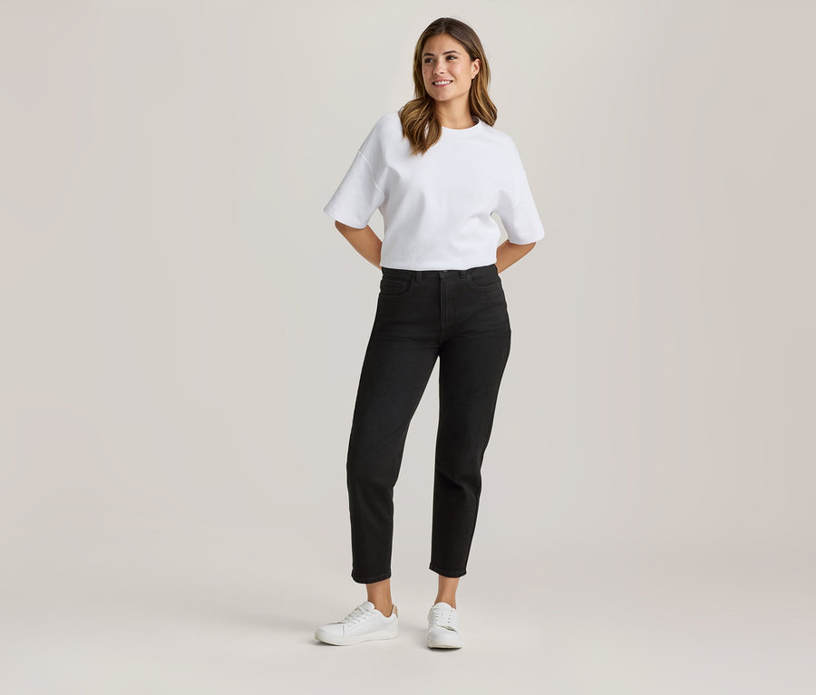 Stehende Frau in weißem Oversize-T-Shirt, schwarzer Cropped-Jeans und weißen Sneakern, mit Händen hinter dem Rücken lächelnd.