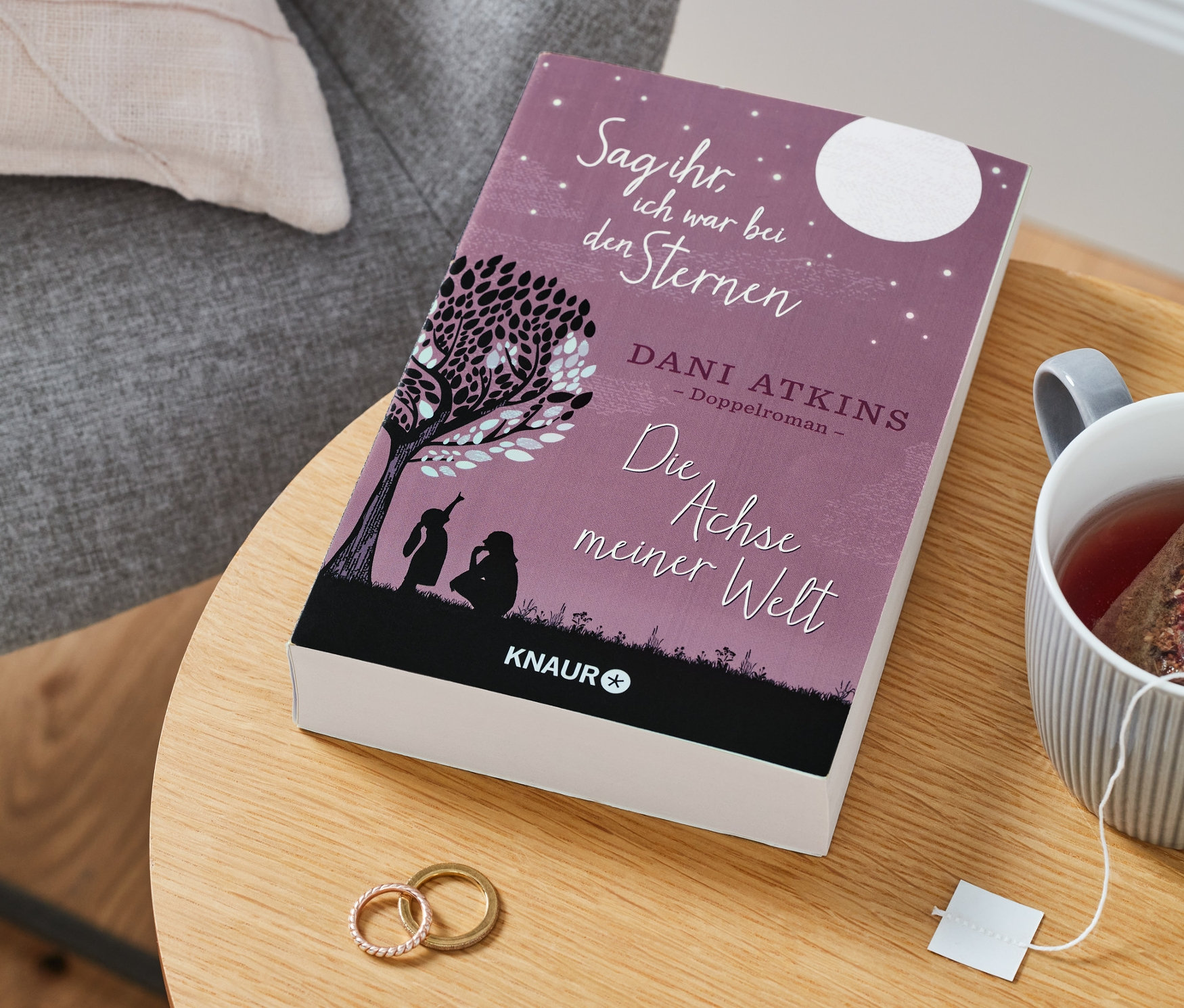 Auf einem Tisch liegen der Doppelroman »Sag ihr, ich war bei den Sternen« und »Die Achse meiner Welt«, eine Tasse Tee und zwei Ringe.