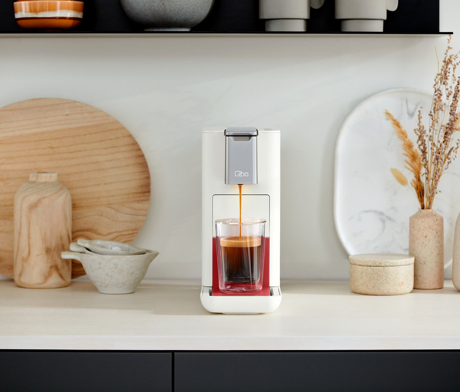 Eine Qbo ESSENTIAL Bright Coral Kaffeemaschine gießt Kaffee in ein Glas. Im Hintergrund befinden sich dekorative Gegenstände und eine Qbo ICONIC Dark Chrome.