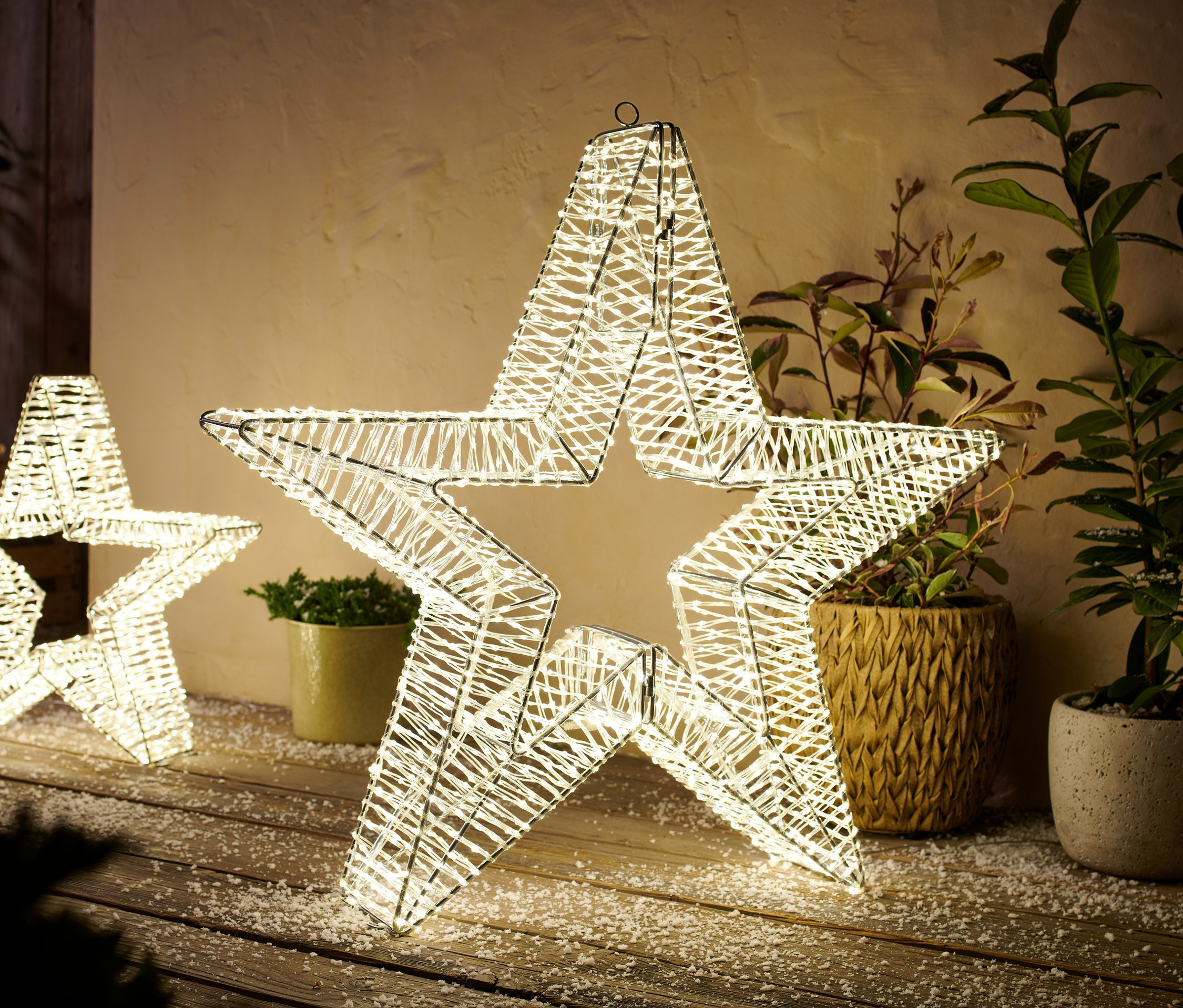 Zwei 3D-LED-Outdoor-Sterne, Größe L, stehen auf einem mit Schnee bedeckten Holzboden. Im Hintergrund befinden sich Pflanzen in Töpfen.
