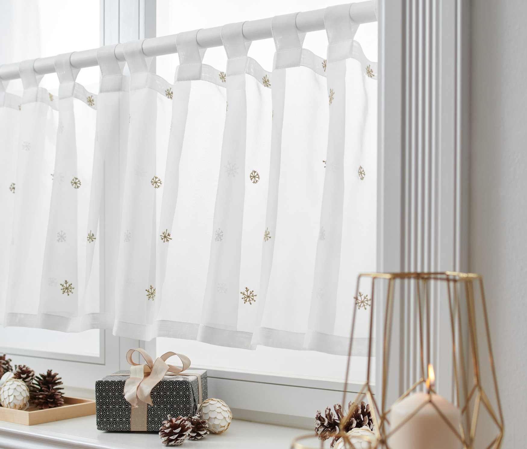 Eine weiße Bistro-Gardine mit Schneeflockenmuster hängt an einem Fenster. Auf dem Fensterbrett liegen eine Geschenkbox und Tannenzapfen.