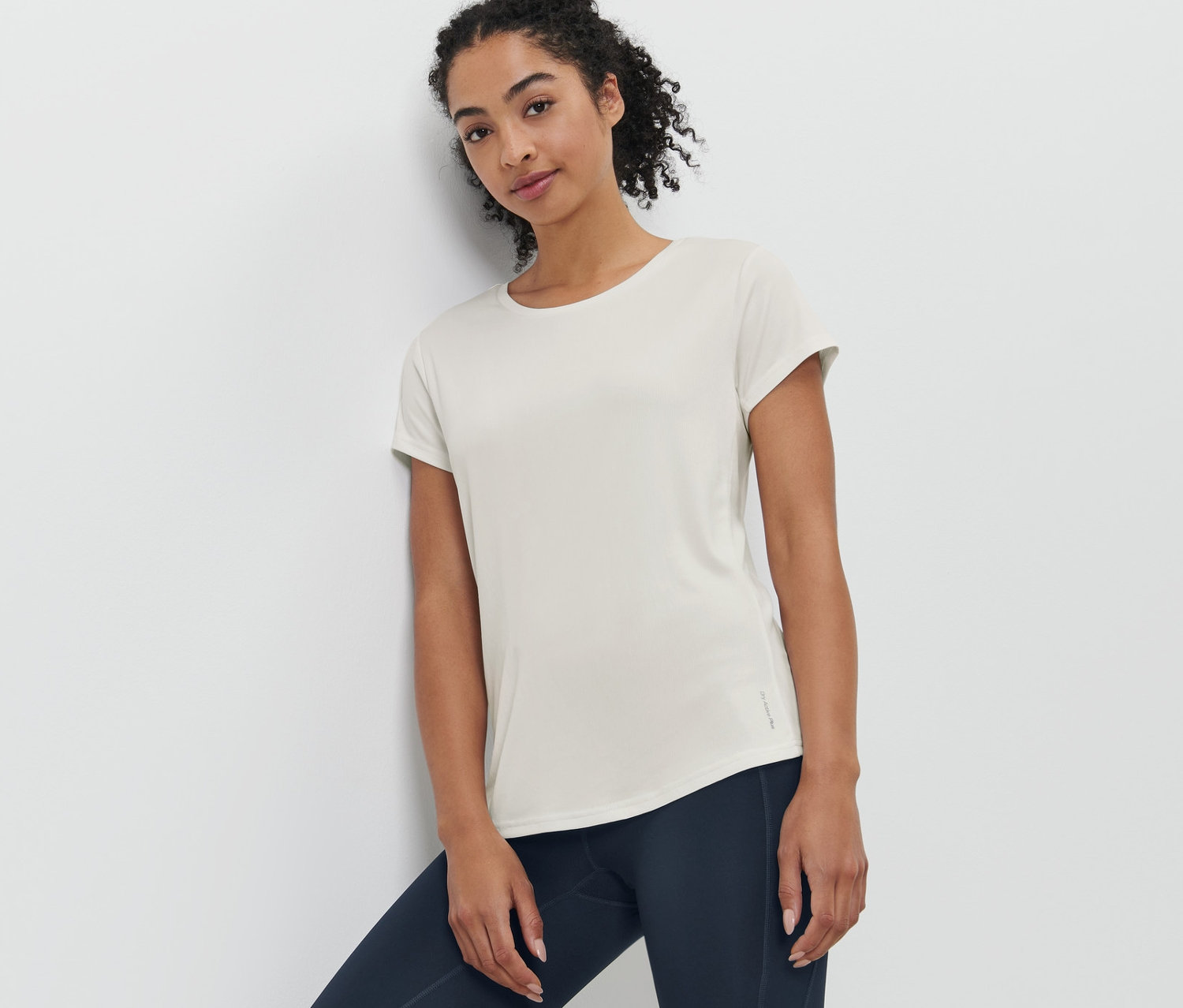 Eine Frau, die sich an eine Wand lehnt, trägt ein offwhite Funktionsshirt und marineblaue Leggings.
