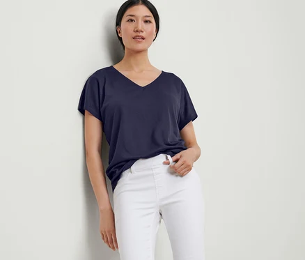 Eine Frau lehnt an einer Wand und trägt ein dunkelblaues, strukturiertes Shirt und eine weiße Hose.