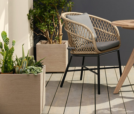 Zwei Dining-Stühle mit Polyrattangeflecht stehen an einem runden Gartentisch. Über dem Tisch hängt eine Solar-Filament-Hängeleuchte. Links steht ein Pflanzkasten auf Rollen.