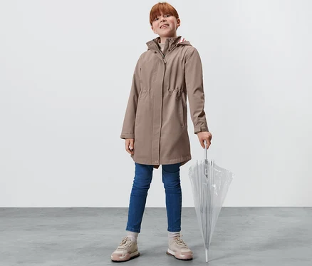 Mädchen steht mit durchsichtigem Regenschirm. Sie trägt einen Kinder-Allwetter-Trenchcoat, Jeans und Turnschuhe.