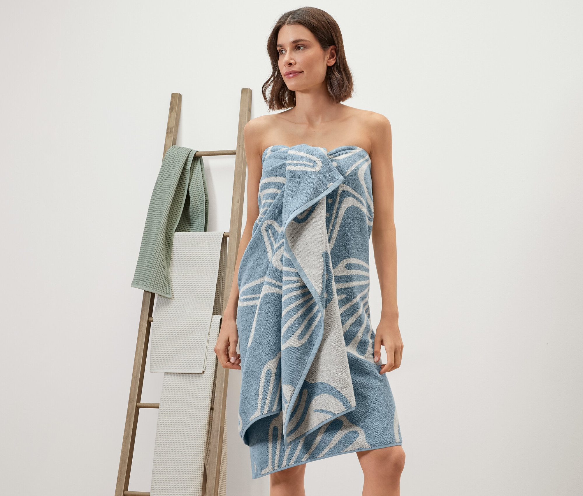 Eine Frau hat ein XL-Badetuch mit Muschel-Muster um sich gewickelt. Im Hintergrund steht eine Holzleiter mit Handtüchern.
