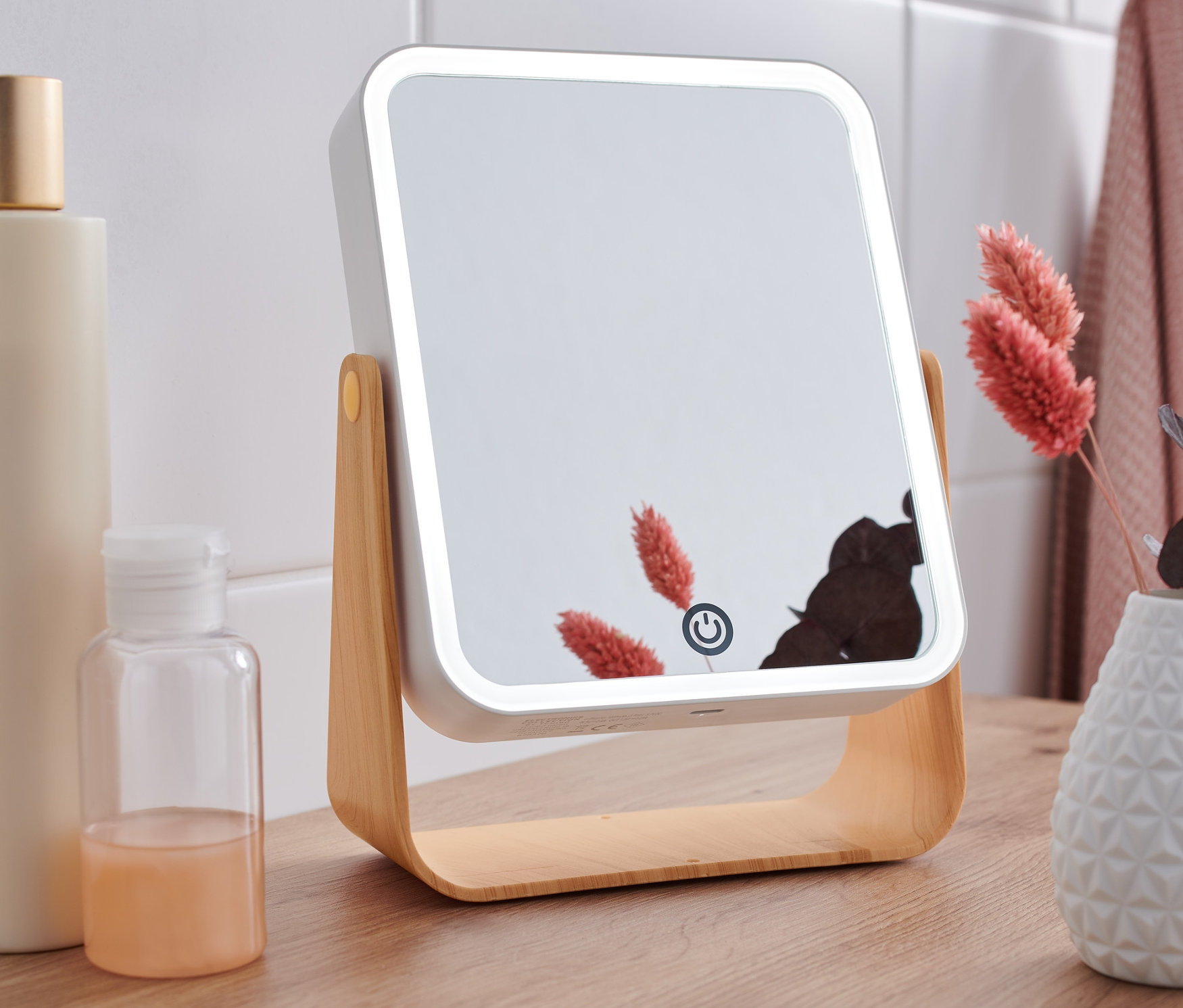 Ein LED-Kosmetikspiegel steht auf einem Holztisch neben Flaschen und einer Vase mit Blumen.