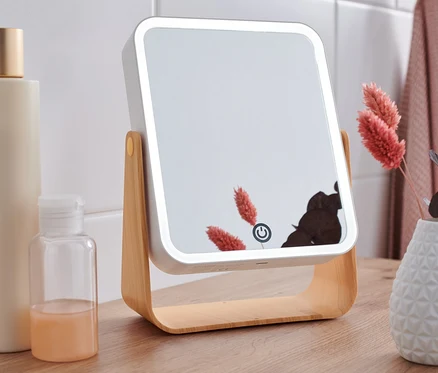 Ein LED-Kosmetikspiegel steht auf einem Holztisch neben Flaschen und einer Vase mit Blumen.