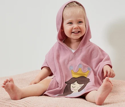 Ein lächelndes Kind sitzt auf einer rosafarbenen Oberfläche und trägt einen Frottierponcho mit Kapuze und einem Prinzessinenmotiv.