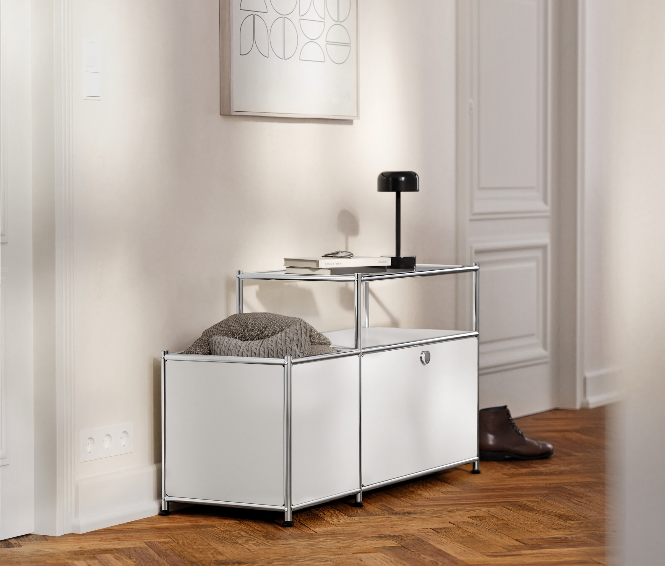 Im Flur steht ein weißes Metall-Lowboard »CN3« mit Klappfach und Glasplatte, auf dem ein Buch und eine Lampe liegen.