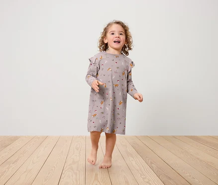 Ein kleines Mädchen in einem Kleid mit Fuchsmuster und Rüschen an den Schultern steht auf einem Holzboden.