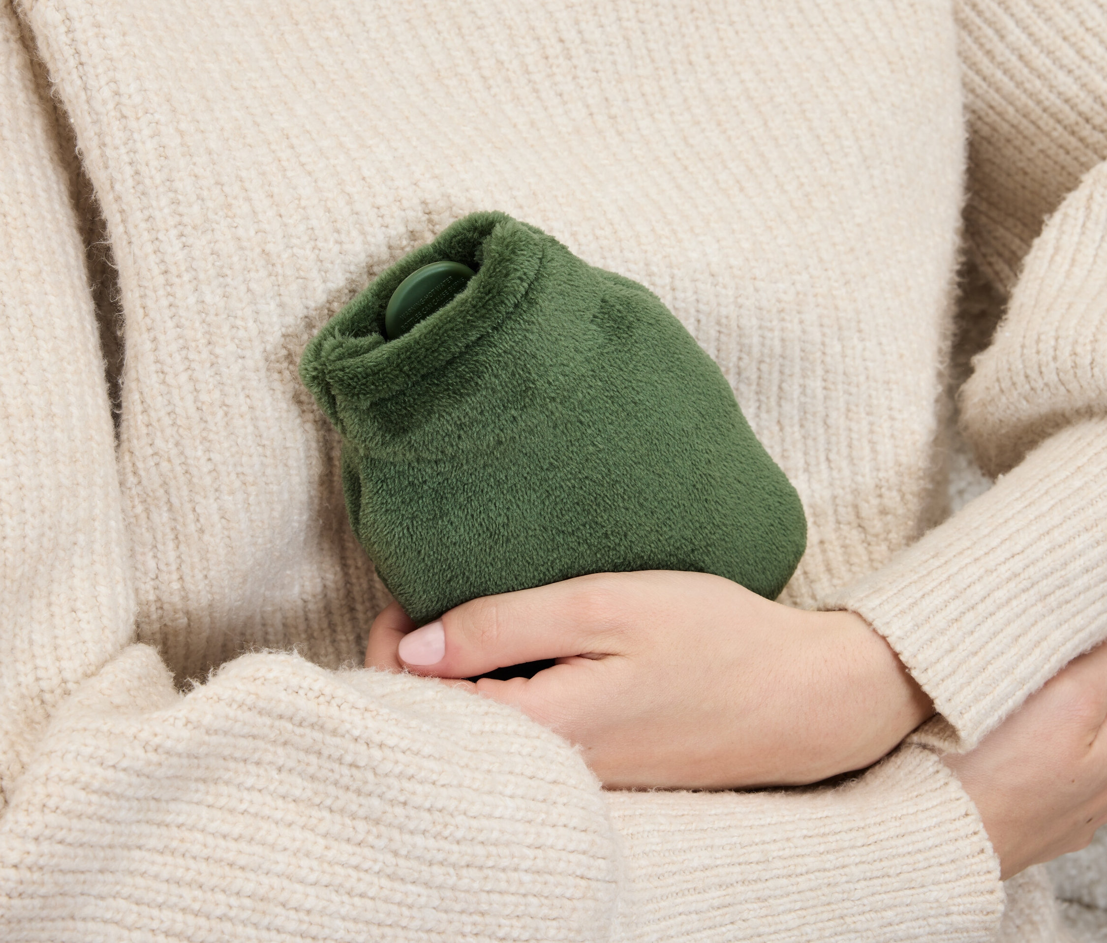 Frau im Pullover hält eine grüne, flauschige Wärmflasche in ihren Händen.
